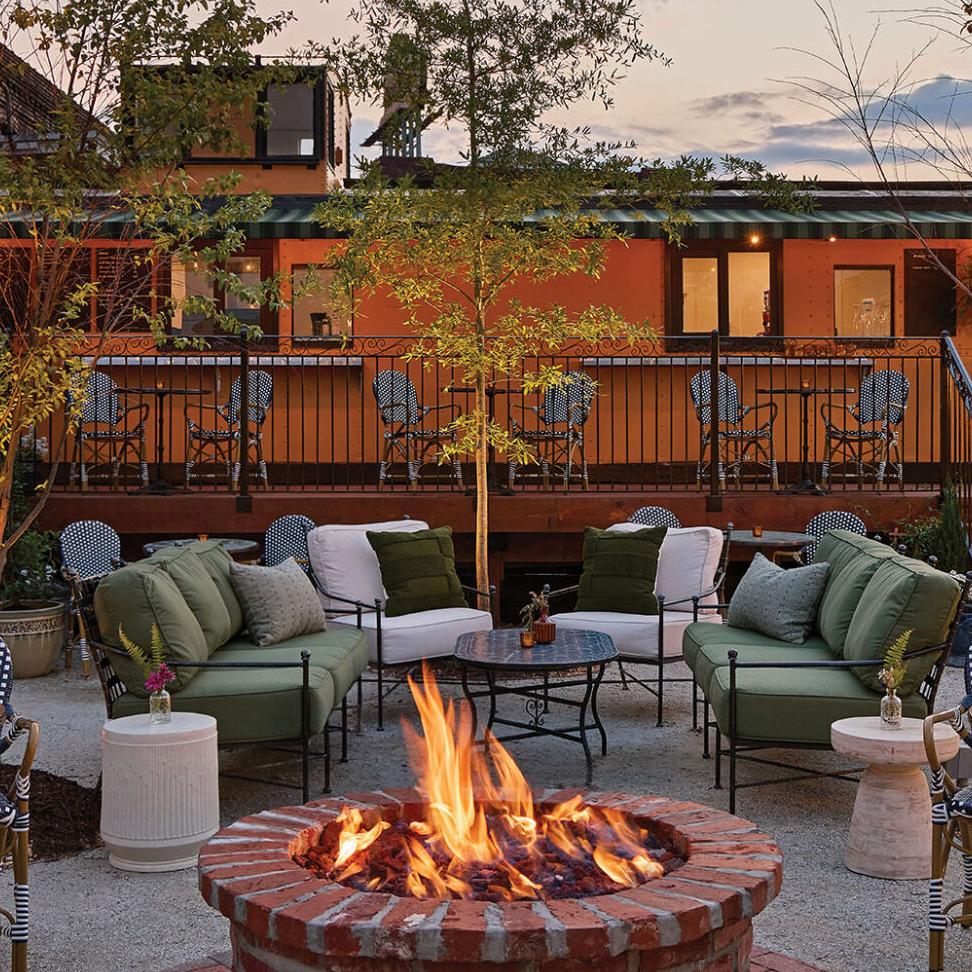 Patio furniture around a circular brick fire pit outside of an orange trailer.
